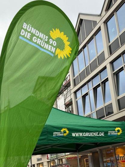 Fotografie einer grünen Beachflag und eines grünen Pavillons der Grünen in Brühl. Beide tragen das Logo von Bündnis 90/ /Die Grünen. Auf dem Pavillon steht außerdem die Webadresse www.gruene.de. Im Hintergrund sieht man eine Gebäudefassade mit vielen Fenstern.