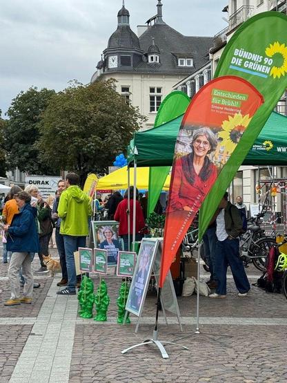 Fotografie eines wahlkampfstands der grünen in Brühl. Vor dem grünen Pavillon stehen zwei Beachflags. Eines mit einem Foto der Bürgermeisterin-Kandidatin Simone Holderried. Diese Beachflag ist rot und trägt den Slogan "Entschieden für Brühl". Die zweite Beachflag ist grün und trägt das Logo von Bündnis 90/Die Grünen mit der Sonnenblume. 
Vor dem Wahlkampfstand stehen grüne Gartenzwerge mit Schildern in der Hand. Außerdem sieht man Passant*innen und Mitgieder*innen der Grünen im Gespräch. Im Hintergrund sieht man das Rathhaus mit seinem Uhrentürmchen