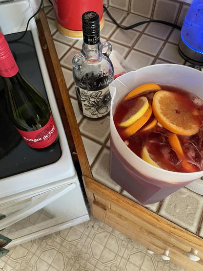 Oranges and lemon slices float in a dark red liquid inside a large jug. There is a red bottle of wine on the stove and a bottle of dark rum on the counter.