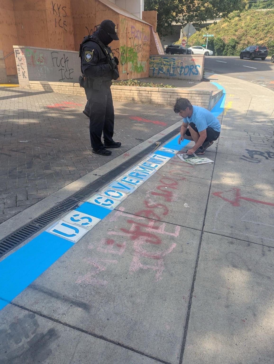 Image shows a DHS agent watching somebody in a blue shirt painting on the ground the words “US goverment”