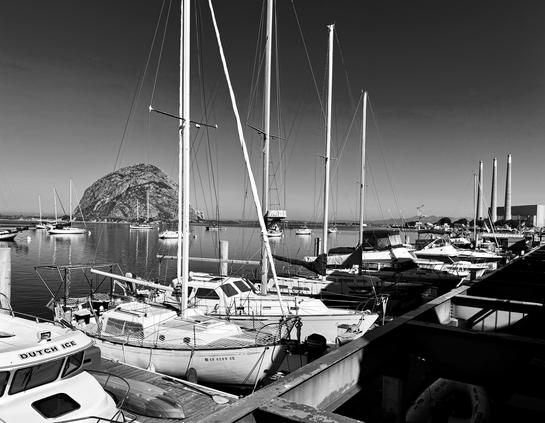 A black and white image of Morro Bay, California. In the foreground are moored boats (mostly sailboats). Across the bay is Morro Rock, and to the far right are the three tall smoke stacks from an old power plant, somewhat iconic, but there is talk of taking them down. Some people would love to see the eyesores come down, others feel they're an iconic part of the area.