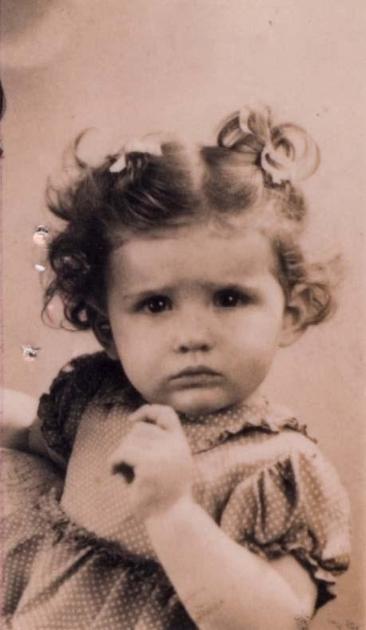 Vintage black-and-white photograph of a young girl with curly hair, wearing a patterned dress, looking directly at the camera.