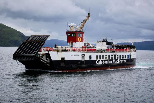 A ferry with 'Caledonian MacBrayne' written on its side moves through calm waters. The deck is visible with lifeboats and railings. Hills and a cloudy sky form the backdrop.