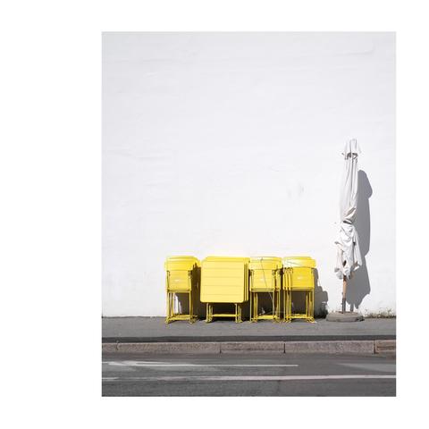 An einer weißen Hauswand stehen zusammengefaltete Stühle und Tische neben einem ebenfalls zusammengeklappten Sonnenschirm. Die Sonne scheint hell.