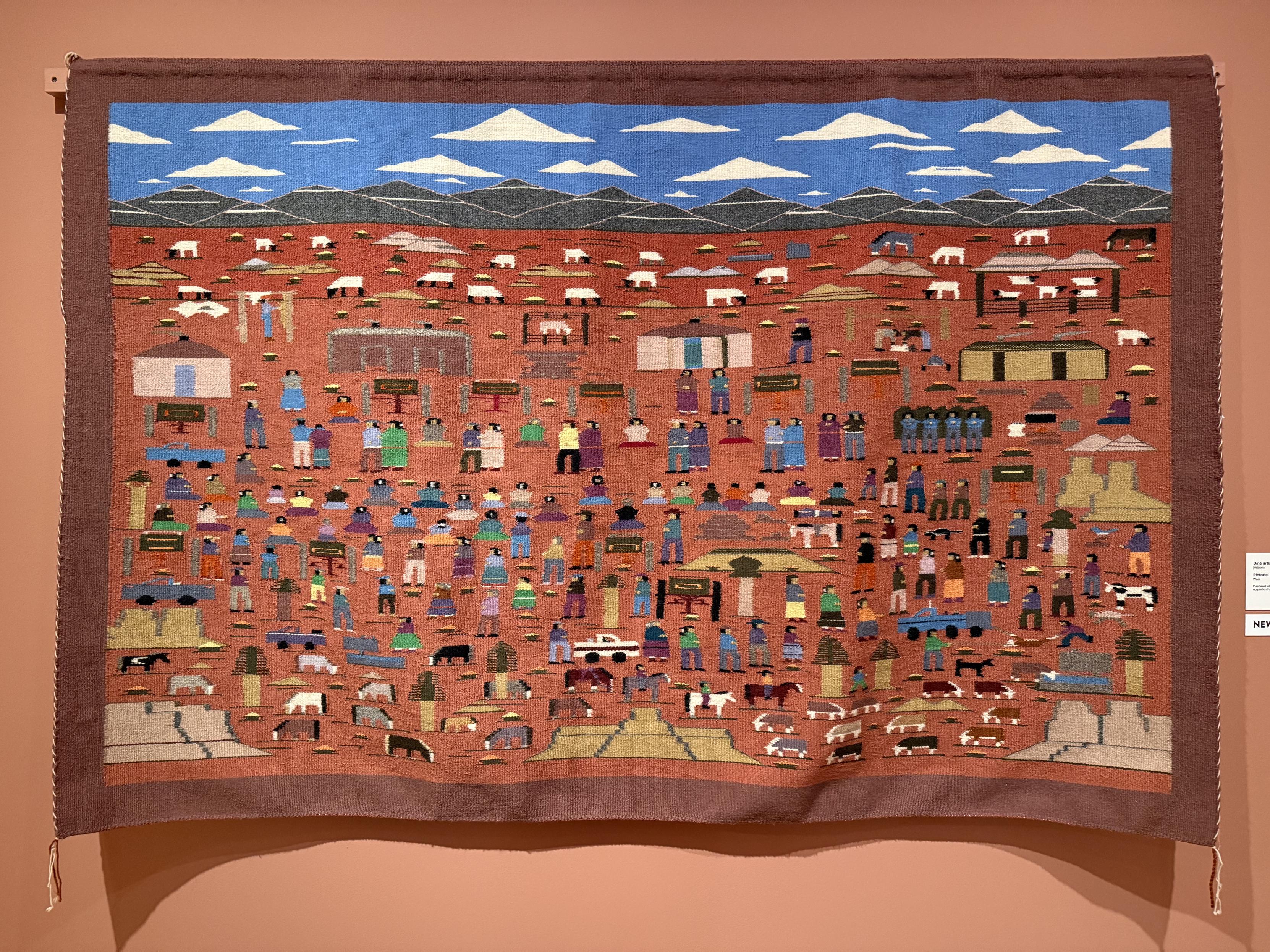 A large pictorial weaving, displaying a busy scene with blue sky and clouds at the top, a thin layer of green mountainous land slightly below that, and a large field of red earth occupying the (majority) space below. All across the earthen scene are various buildings, people, animals, and implements of daily life.