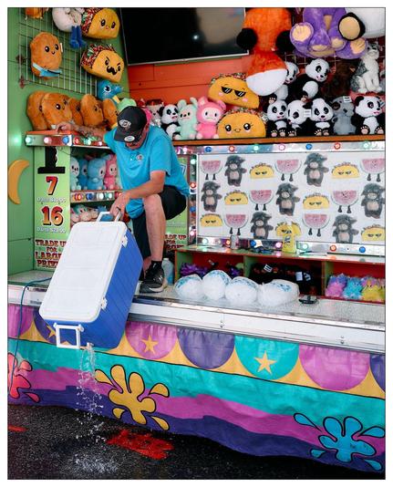 Man in a blue shirt and cap, seated at a carnival game stall, emptying water from a cooler. The stall is decorated with colorful artwork and arrayed with various plush toys like emojis and pandas as prizes. Visible game targets feature cartoon sheep faces, and the booth has a vibrant, multicolored base with star and flower motifs.