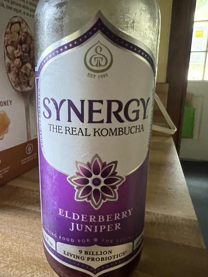 The image shows a bottle of Synergy Kombucha placed on a wooden surface. The label is predominantly purple and white with decorative accents. At the top of the label is the Synergy logo, including “EST 1995.”

The large text in the middle reads:
“SYNERGY – THE REAL KOMBUCHA.”

Below that, it specifies the flavor:
“ELDERBERRY JUNIPER” with a stylized flower-like design above it.

At the bottom, the label highlights:
“9 BILLION LIVING PROBIOTICS!”

The bottle is chilled, with condensation visible on the glass. In the background, part of a cereal box and a door can also be seen.