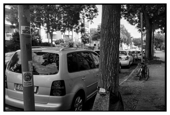 Black and white photo in landscape orientation. A row of parked cars at the side of the road. On a sign post a sticker from Fridays For Future Berlin: RADWEG GESUCHT (looking for the cycle path).