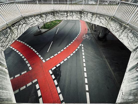 A road intersection viewed from above, featuring a distinctive red X-shaped bike lane. The bike lane is bordered by dashed white lines. A white bicycle symbol is visible on the pavement, indicating a cyclist route. The intersection is surrounded by a concrete bridge with white railings, providing an elevated perspective. The area has visible markings and weathered textures on the concrete surfaces.