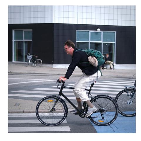 Ein Radfahrer gekleidet wie im Post beschrieben. Er trägt außerdem noch einen lässigen Rucksack mit dem er ein Sacko zwischen Rucksack und Rücken gelagert hat.