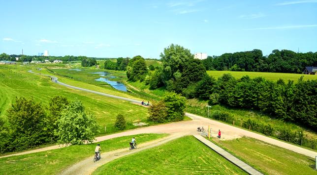 Cyclists and pedestrians travel along a scenic cycle route, winding beside a river through nature, surrounded by trees and grassy fields under a clear blue sky.

Radfahrer und Fussgaenger fahren auf einem malerischen Radweg, der sich neben einem Fluss durch die Natur schlaengelt, umgeben von Baeumen und Grasfeldern unter einem klaren blauen Himmel.