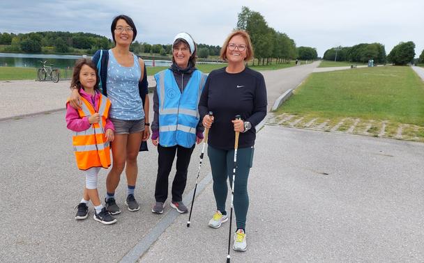 In the paths of a park, by a lake, four people pose for a photo. First, we see a little girl of about seven years old, dressed in sportswear, wearing the tail walker's high-visibility orange vest. Next to her is her mother, also dressed in sportswear, holding her by the shoulder. On the right, a woman is wearing the sky blue high-visibility jacket of the park walker. She is wearing a rain jacket, trousers and trainers. Finally, another woman, also in sportswear, is holding a walking stick in each hand.