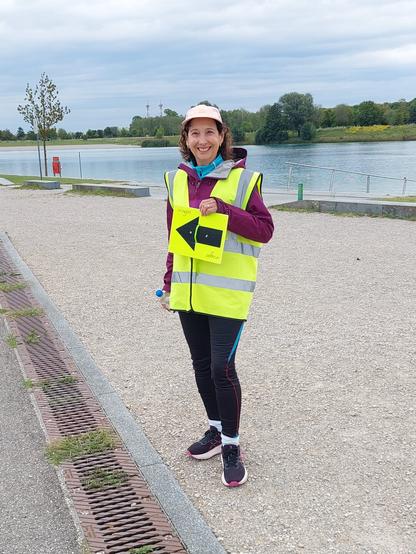 With the lake in the background, a smiling woman holds a black arrow on a yellow background in her left hand, pointing to the left. The woman is wearing a high-visibility yellow parkrun volunteer jacket. She is wearing a cap, two jackets, one of which is a rain jacket, running leggings and running shoes. She is holding a bottle of water in her right hand. The sky is grey.