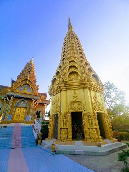 Photo shows to temp two temple buildings in yellow gold painting and decorations. In front there is a stupa-like tower covered in light yellow gold, with six open gates at ground level. Inside there are altars of holy figures with offerings of light and incense. Over the gates there are bas-reliefs with variations of motifs of the life of the first Buddha. The heavily arched roof of the spire each have detailed carvings matching the bas reliefs. To the left there is a pagoda in yellow, orange and gold with intrinsic details in the decorations covering the pagoda up to the seven-level tower. The locked double door panes to the pagoda are in gold, with two visible guards engraved on each door. The stair up to the pagoda is painted blue with a pink/purple center stripe. It is a sunny day with clear blue sky. To the right there is a cluster of trees. Cambodia is as safe as ever, and welcomes visitors.
