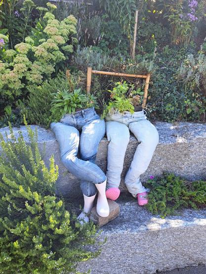 Zwei Bluejeans ausgefüllt, als wären Beine drin. Aus der Taille wachsen Pflanzen. "Füsse" sind Gummistiefel und pinke Crocs