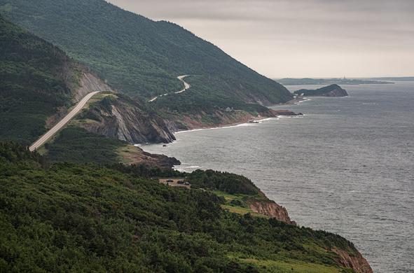On the left, a winding road along a steep coastal mountainside on a cloudy day. Calm waters outline the coast and the horizon in the far distance.