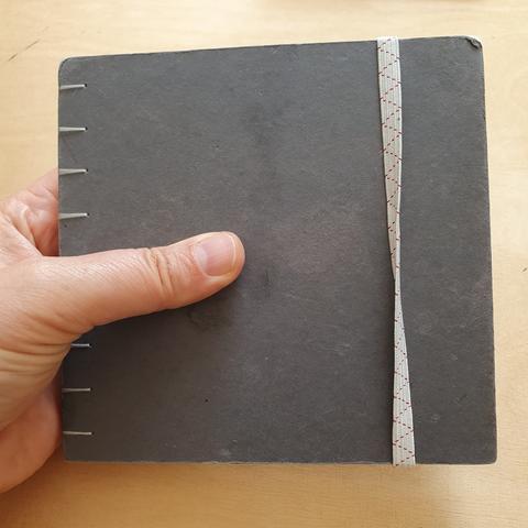 Foto vom dunkelgrauen quadratischen Skizzenbuch, das von einer Hand gehalten wird.