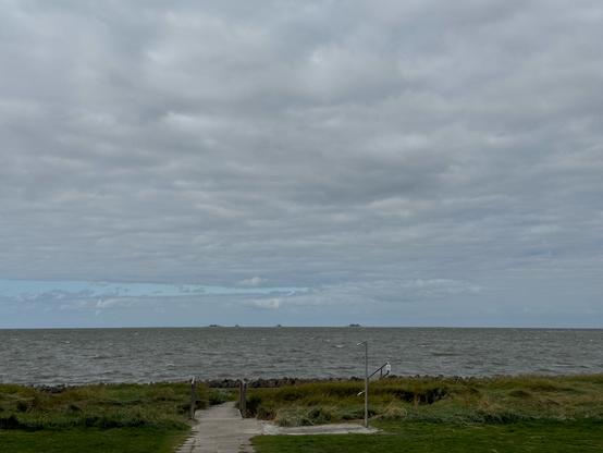 In the foreground a paved walkway leads through a grassy area past an open air shower to the North Sea. 
Waves break in the sea and the sky is mostly overcast.