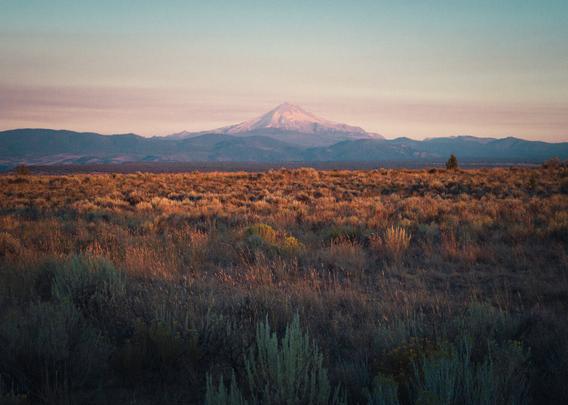￼a glaciated mountain Peak rises above a vast sandy landscape filled with small desert plants, clear skies laced with ribbons of smoke from a wildfires only miles away