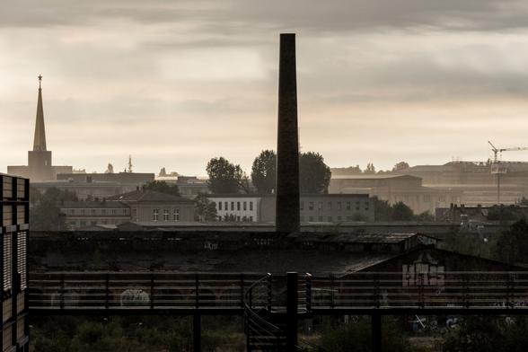 DE: Eine urbane Skyline mit Blick nach Osten. Wolken und leichter Nebel korrespondieren mit den dunkel gehaltenen Gebäuden. Zentral erhebt sich ein Industrieschornstein von einem alten Ringlokschuppen. Der Russische Pavillon auf dem alten Messegelände erkennt man gut an seinem spitzen Dach mit dem (roten) Stern an der Spitze.

EN: An urban skyline with a view to the east. Clouds and light fog correspond with the dark buildings. An industrial chimney rises centrally from an old roundhouse. The Russian Pavilion on the old exhibition grounds is easily recognisable by its pointed roof with the (red) star at the top.