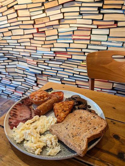 Full English breakfast (eggs, bacon, pork sausage, hash brown, mushrooms & toast) on a table, a wall of books in the background