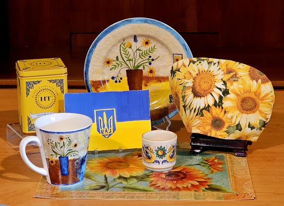 A boldy yellow and blue tea tableau in homage to the spirit of Ukraine. Upon a sunflower pattern placemat are a sunflower tea cozy, sunflower cup, plate, and mug, a handpainted Ukrainian Trident flag, and Yellow and Blue Tea from Harney and Sons, a chamomile and lavender herbal infusion from which the sale proceeds yield a contribution to Ukrainian relief.