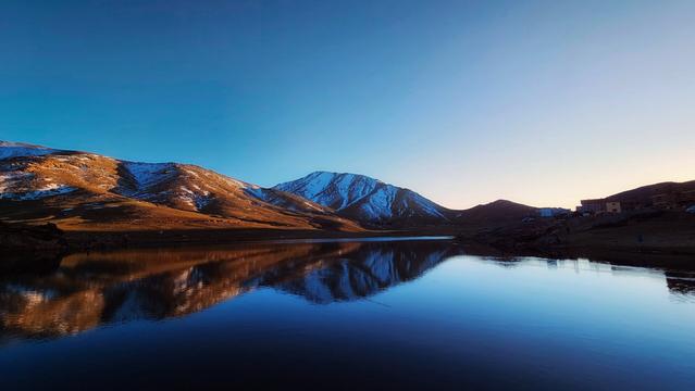 A panoramic view of a calm, reflective lake at sunset, with a range of mountains in the background. The snow-capped peaks of the mountains are illuminated by the warm, golden light of the setting sun, contrasting with the cool blue tones of the sky and the water. The mountains are reflected almost perfectly in the still surface of the lake, creating a stunning symmetrical image. A few buildings are visible on the right bank of the lake.