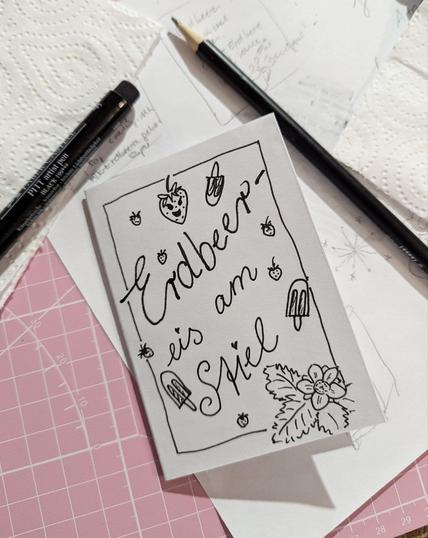 Rosa Schneidematte. darauf Papier, Stifte und ein Mini-Zine. Auf dem Deckblatt steht "Erdbeereis am Stiel" in schwarzer Schreibschrift. Runtherum sind mit dem gleichen Stift kleine Erdbeeren, Eis am Stiel und eine Erdbeerblüte mit Blättern gemalt.