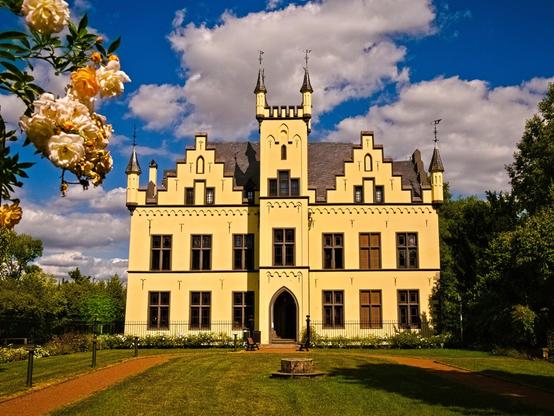 This neo-Gothic and neo-Renaissance mansion is a two-story plaster building with an almost square floor plan and a tower, with the building sections extending to the left and right. The façade is yellow. The inner courtyard is largely occupied by an ornamental garden. The sun is shining. And there are a few roses on the upper left side of the photo.
