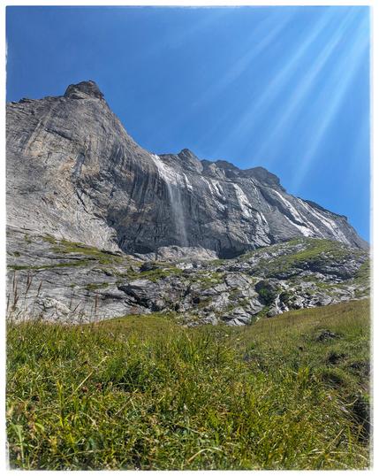 View step up from a hiking path, across a meadow, to a rocky cliff with a small waterfall.