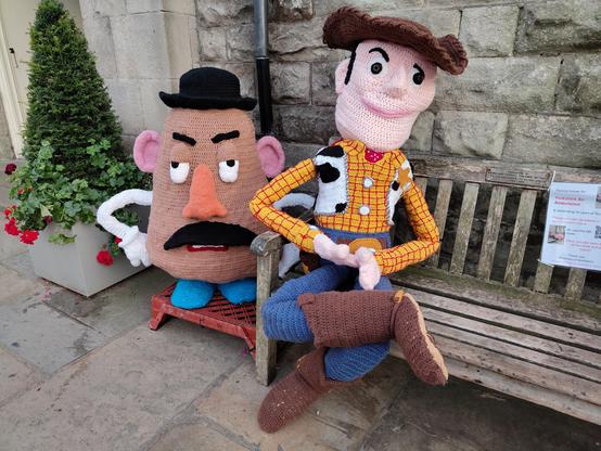 Potato steht neben einer Bank, auf der Woody sitzt. Woody ist menschengroß, Potato dazu passend.