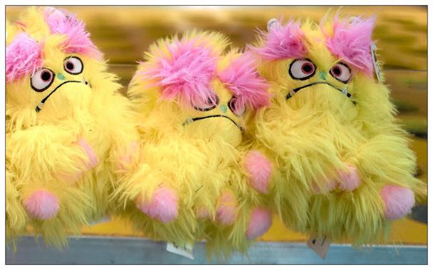 Three yellow and pink plush toys with exaggerated, cartoonish eyes and fluffy bodies hang against a blurred yellow background. These toys feature pink tufts on top, googly eyes with prominent lashes, and a dismayed expression drawn on their faces. Each has pink spherical cheeks and a body predominantly yellow with a hint of pink near the top.