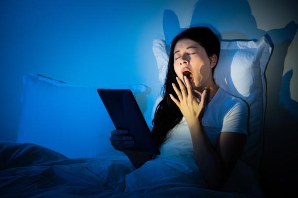 young asian woman in bed rreading a tablet and yawning, blue tones - deposit photos