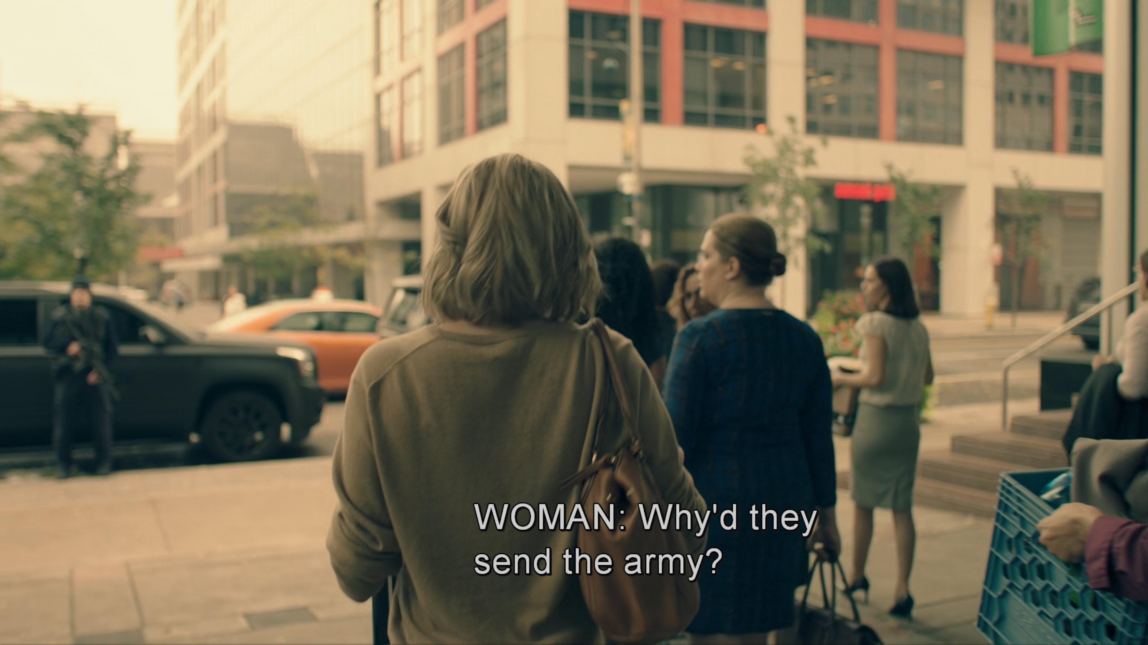 Screenshot from The Handmaid's Tale TV series. June, a blonde woman walks with other women as they exit their office building after being fired. Beyond them are cars and modern multi-story buildings. Other dismissed female employees can be seen in the background. The subtitle shows a woman asking "Why'd they send the army?" This scene occurs immediately after June and all the women at her office are fired due to a new law prohibiting women from holding jobs, as they notice the unusual military presence on the streets.