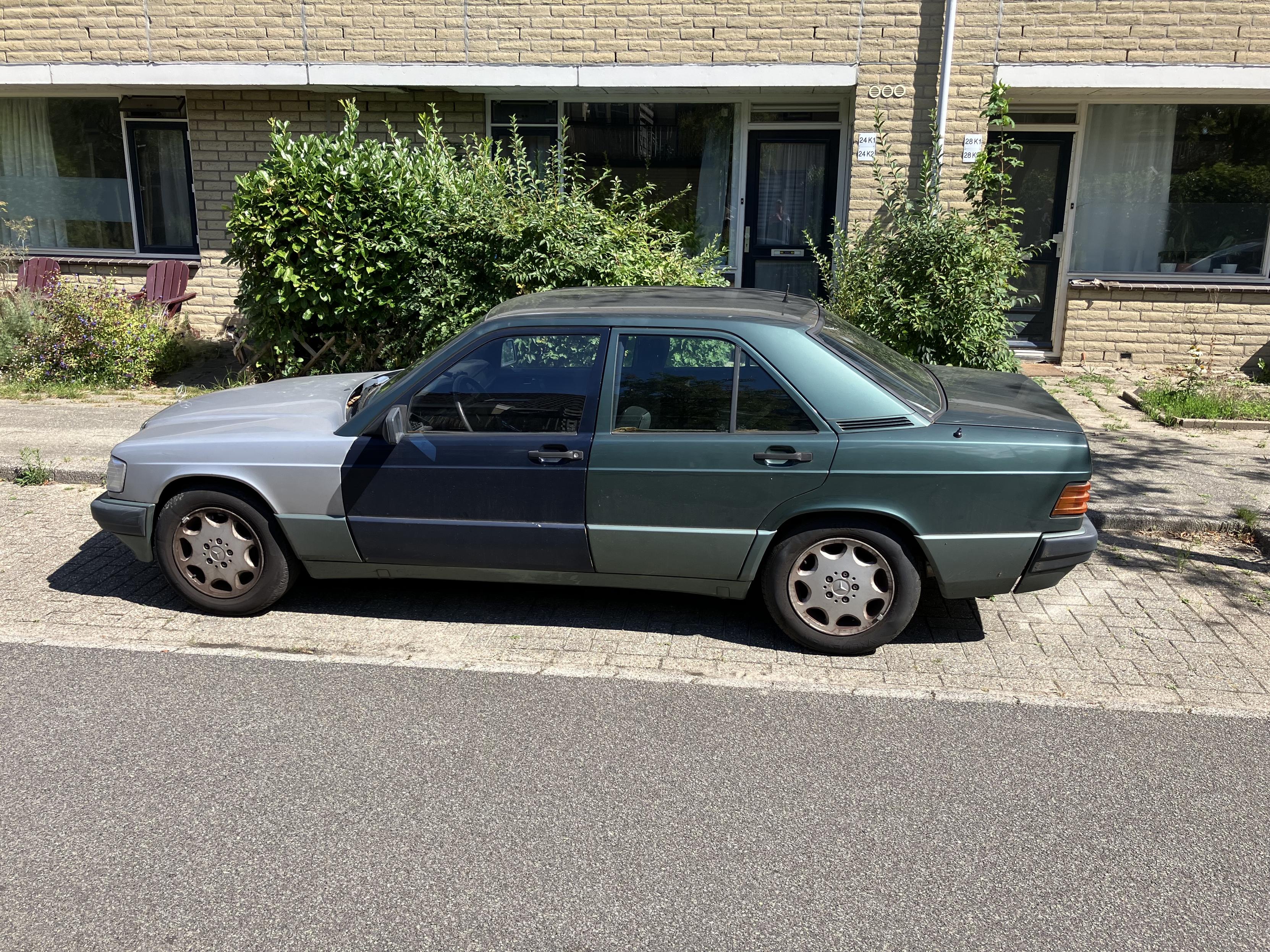 An old Mercedes with a silver grey front, navy driver side door and green roof and back half. It’s parked in front of a light brick building on a sunny day.