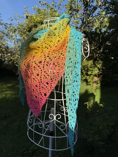 Crocheted turquoise - yellow - pink shawl draped over the shoulders of a mannequin. Green bushes in the background.