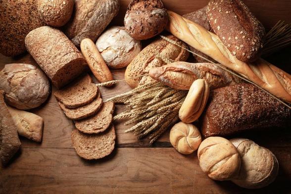 Panes y rebanadas de pan de varios tipos, incluyendo baguettes, panecillos y pan integral, sobre una superficie de madera con tallos de trigo. Fotografía artística.
