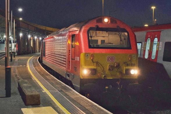 A heavy diesel Engine in red and yellow livery is working the train and waiting for departure at Holyhead station. It's still dark and headlights switched on.