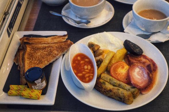 Picture of the full Welsh breakfast. It'@ 2 glamorgan sausages, half a grilled tomato, slice of black pudding one fried egg, two slices of ham, two hash browns accompanied by 4 slices of toast with butter on another plate. Two cups of tea in the background.