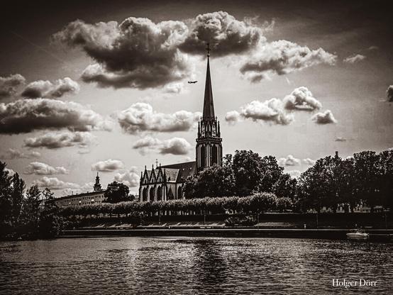 Schwarz-Weiß-Foto der Dreikönigskirche in Frankfurt am Main mit markantem Turm, Wolkenhimmel und Main im Vordergrund.