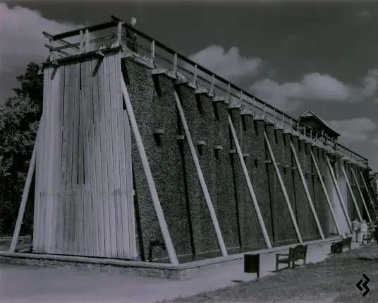 Black and white photo of graduation tower of a salina. The graduation tower is a wall like structure, about 10 meters high, that is stuffed with blackthorn twigs. Water is pumped up an trickles down through the twigs, this causes some water to evaporate and increases the salt concentration in the water. On the foot of the building are basins to catch the water to be pumped up again or extracted for further processing. The building is located in a park with footpaths and benches around it. On the roof is a statue of a man looking down. There is a tree in the background and the sky has some prominent clouds, enhanced by the usage of a red filter for the photograph
