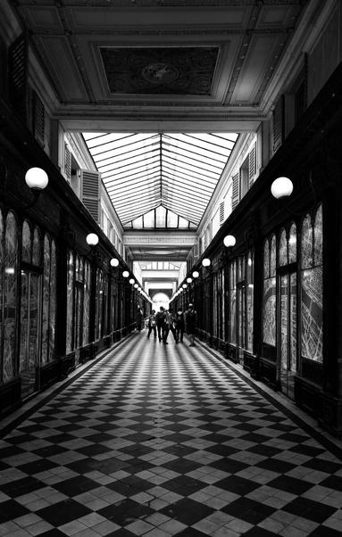 A black and white photograph of an ornate, symmetrical covered walkway or shopping arcade. The ceiling is a mix of glass panelling and decorative plasterwork, with symmetrical rows of decorative streetlights on both sides. The tiled floor has a distinct black and white checkered pattern, which creates a strong sense of depth and perspective, leading the eye towards a small group of people in the distance. The walls are lined with windows and what appear to be maps or blueprints.