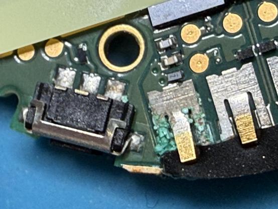 A close-up image of a circuit board featuring various electronic components, including a black connector, gold contacts, and solder points, with traces of corrosion visible. The background is a blue surface.