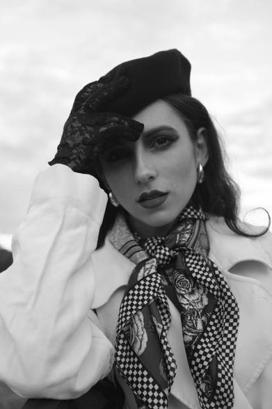 Black and white portrait of a person wearing a dark beret, lace glove, and hoop earrings. They have long, dark hair and are dressed in a light coat with a patterned scarf featuring checkered and floral designs. The backdrop is a cloudy sky.