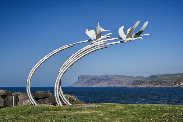 A metal sculpture on the Ballycastle seafront depicting the Children of Lir. Three stylized white swans are shown in flight, mounted on two long, arcing silver metal poles that curve upwards and to the right. The sculpture is set against a clear blue sky, with a rocky shoreline and the sea in the foreground. In the distance, the prominent headland of Fair Head can be seen.