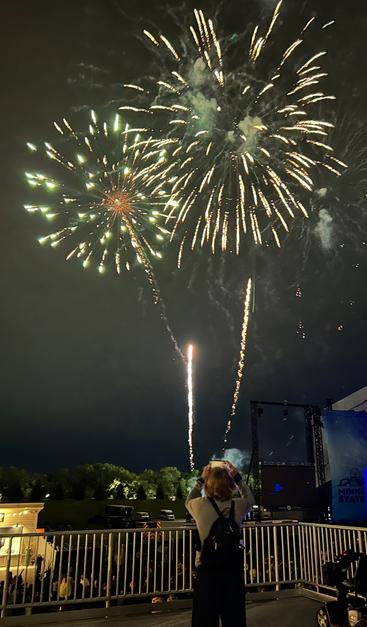 A night sky is lit up by several colorful fireworks, with two having just burst and illuminating the sky, and two other flares streaking upward, having just been shot off from the ground. A person has their back to the viewer and is in the foreground looking upward and holding what looks a smartphone, taking a photo of the same fireworks