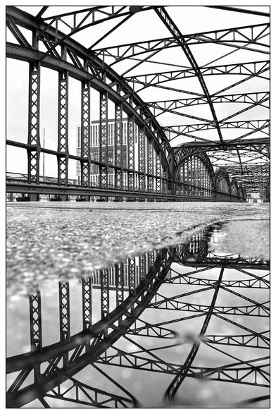 Das Bild zeigt eine Brücke aus Stahl, aufgenommen in Schwarz-Weiß. Die Konstruktion besteht aus mehreren Bögen und Querstreben, die sich über die gesamte Länge der Brücke erstrecken. Die Perspektive ist sehr niedrig gewählt, fast auf Straßenniveau, wodurch die architektonischen Details besonders betont werden. Im Vordergrund befindet sich eine Pfütze auf der Fahrbahn, die eine klare Spiegelung der Brücke zeigt und so die Struktur doppelt erscheinen lässt. Im Hintergrund sind einige Gebäude zu erkennen, die jedoch durch die dominierende Brückenkonstruktion in den Hintergrund treten. Die Gesamtwirkung ist grafisch und kontrastreich, mit einem Fokus auf Linienführung und Symmetrie.
