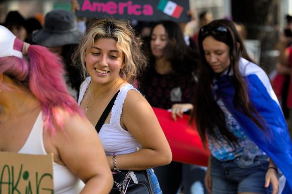 A group of people dancing to ranchero music prior to a march in support of immigrant rights. A tight crop cuts off a lot of dancers except for 6; only 1 is in focus as the subject. The subject is a woman with blond highlights wearing a white top; she is partially obscured by another dancer holding a sign. She's smiling as she dances.