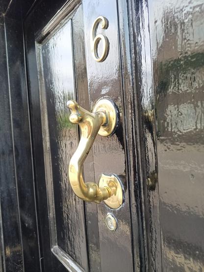 an einer schwarzen Tür ist in Kopfhöhe ein golden farbener Türklopfer angebracht.
Die Form ist wie ein S, wobei der Teil der gegen die Tür klopft, der Hals und Kopf eines Menschen ähnlichen Wesens ist