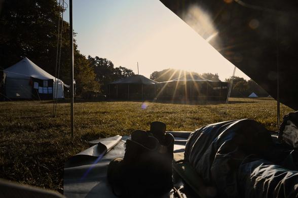 Blick aus einem Zelt kurz nach Sonnenaufgang. Im Vordergrund Wanderschuhe und ein Schlafsack, dahinter Wiese. Im Hintergrund weitere Jurten und die Sonne.